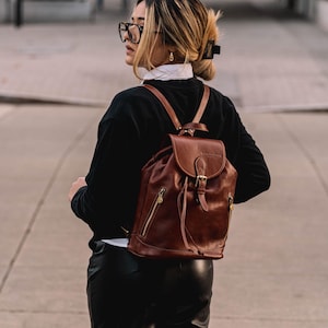 Puede incluir: Una mochila de cuero marrón con cierre de solapa, hebilla y cordón. La mochila tiene un asa superior y correas ajustables para los hombros. Cuenta con un bolsillo frontal con cremallera y herrajes dorados. La bolsa se lleva en la espalda.
