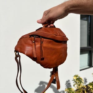 A close-up of a brown Italian leather belt bag (bum bag) being held by hand, showcasing its soft texture, quality craftsmanship, and sleek design.