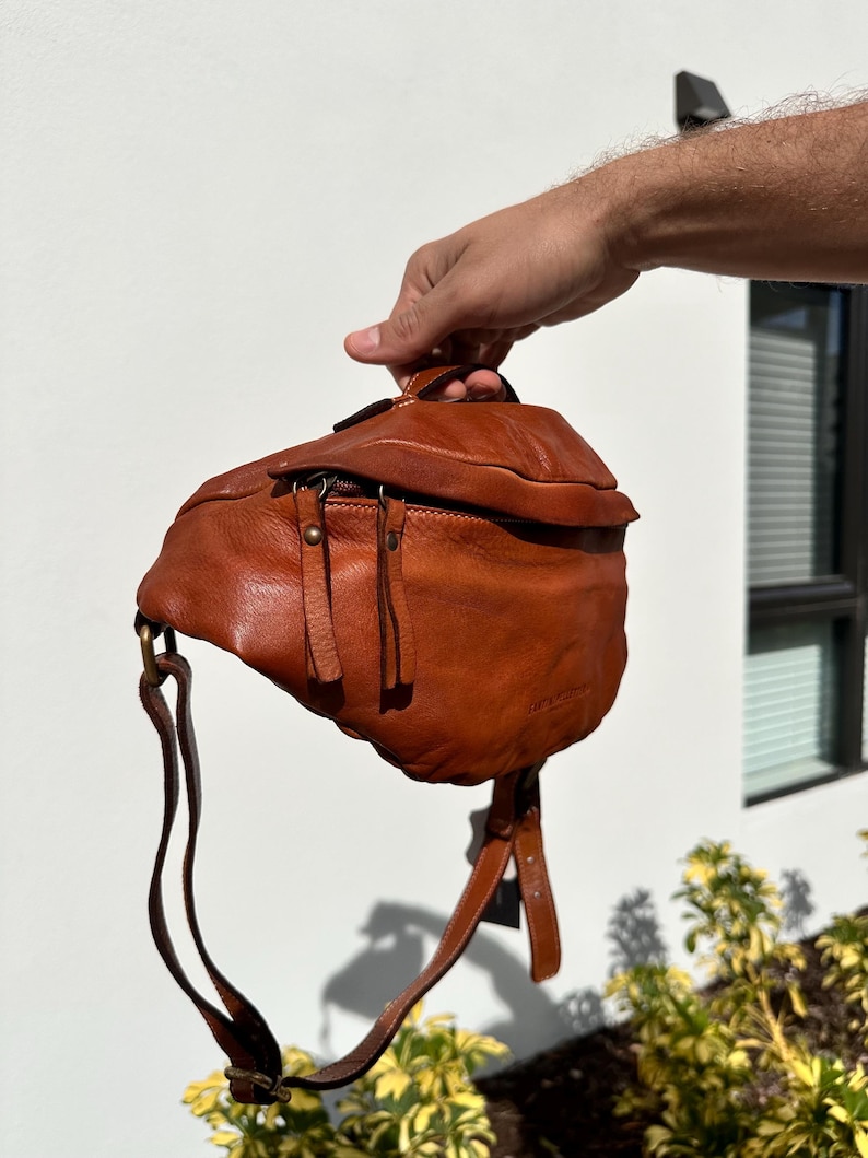 A close-up of a brown Italian leather belt bag (bum bag) being held by hand, showcasing its soft texture, quality craftsmanship, and sleek design.