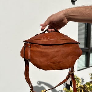 A close-up of a brown Italian leather belt bag (bum bag) being held by hand, showcasing its soft texture, quality craftsmanship, and sleek design.