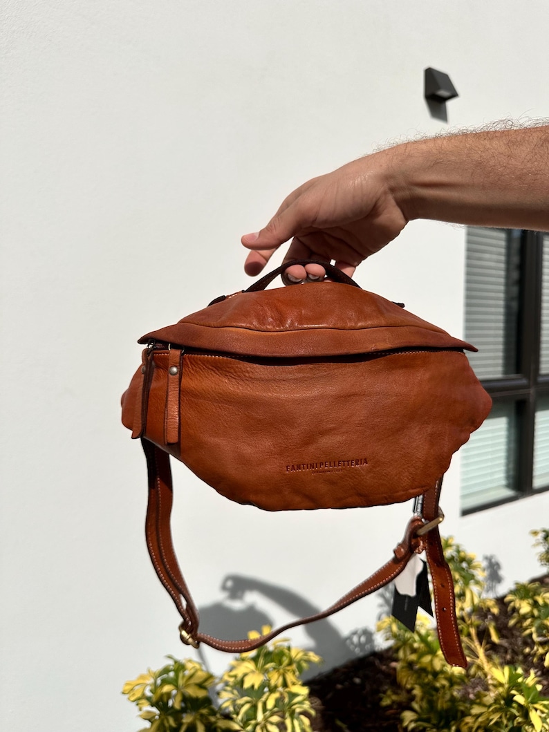 A close-up of a brown Italian leather belt bag (bum bag) being held by hand, showcasing its soft texture, quality craftsmanship, and sleek design.