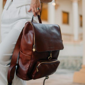 May include: A brown leather backpack with gold accents and zippers. The backpack has a flap closure and a front pocket.