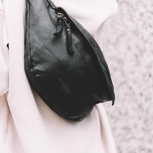 Black leather hip bag draped over a white outfit. This modern waist bag offers a sleek, minimalist look suitable for any outfit. A luxurious yet practical leather travel bag for those who value both style and convenience
