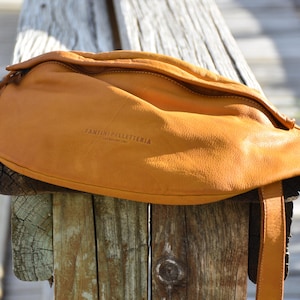 Close-up of a tan leather fanny pack resting on a weathered wooden post, showcasing its natural texture and craftsmanship, perfect for adding a pop of color to any outfit.