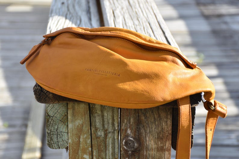 Close-up of a tan leather fanny pack resting on a weathered wooden post, showcasing its natural texture and craftsmanship, perfect for adding a pop of color to any outfit.
