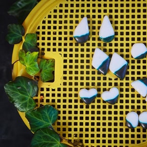 Può includere: Assortimento di piastrelle in ceramica di varie forme, tra cui cuori, diamanti e lacrime. Le piastrelle sono dipinte in uno schema di colori bianco e nero con un accento turchese.