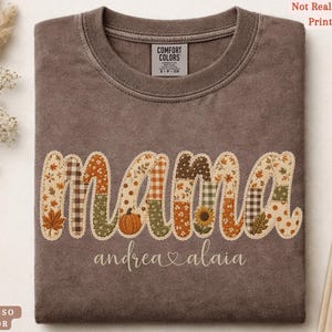 May include: An espresso-colored crewneck t-shirt featuring the word mama in a patchwork-style print with autumn-themed patterns including pumpkins, sunflowers, and leaves. Below the word mama, the names andrea and alaia are printed in white script.