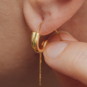 May include: A gold hoop earring with a delicate chain hanging from it. The earring is being put on by a person.