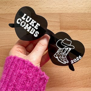 May include: Black heart-shaped sunglasses with the text "LUKE COMBS" in white on one lens. The other lens features a cowboy hat and boots graphic, with the number "2026" in white. The sunglasses are held up by a person wearing a pink jumper.