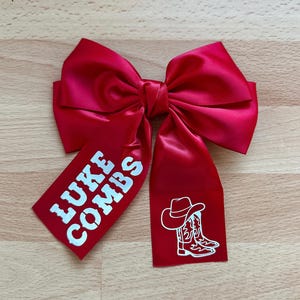 May include: A vibrant red satin bow with white text reading "LUKE COMBS" on one tail. The other tail displays a white graphic of a cowboy hat and boots. This bow is likely for hair or gift wrapping.