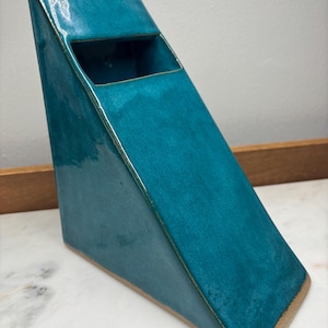 May include: A teal ceramic wall pocket with a glossy finish and a triangular, wedge-shaped profile. The object features a flat, angled front surface with a rectangular opening near the top, and a flat base, showcasing a smooth, glazed exterior.