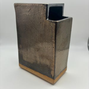 May include: A rectangular ceramic vase with a dark brown glaze and a light brown rim. The vase has a unique, asymmetrical opening at the top.