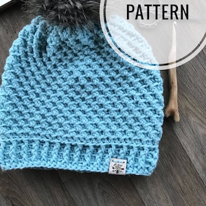 Puede incluir: Un gorro de punto azul claro con un pompón gris esponjoso en la parte superior. El gorro tiene un patrón de punto texturizado y una etiqueta blanca con el texto "Crochet Love" en él.