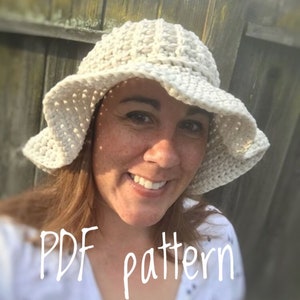Può includere: Un cappello da sole bianco all'uncinetto con un'ampia tesa e un design a uncinetto. Il cappello è indossato da una persona con i capelli castani e una camicia bianca. Il testo "PDF pattern" è visibile nell'immagine.