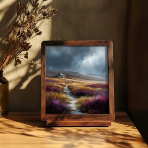 May include: A framed landscape print depicts a path leading to a small house on a hill under a cloudy sky. The path winds through fields of purple and gold flowers. The frame is made of dark wood.