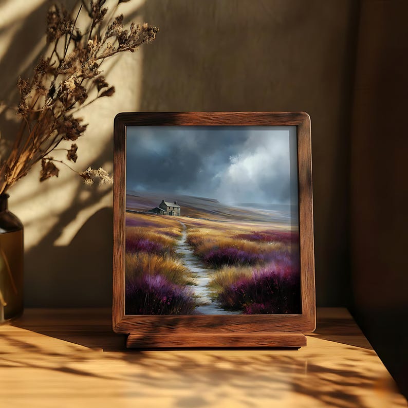 May include: A framed landscape print depicts a path leading to a small house on a hill under a cloudy sky. The path winds through fields of purple and gold flowers. The frame is made of dark wood.