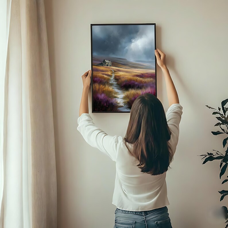 May include: A framed landscape art print depicts a path leading to a small house under a dramatic sky. The artwork features a color palette of purple, gold, and gray, with a black frame. The print is being held up against a white wall.