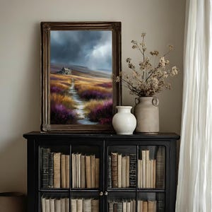 May include: A framed landscape painting depicts a path leading to a small house under a stormy sky. The path winds through fields of purple and gold flowers. A white vase and a rustic ceramic vase with dried flowers sit on a black cabinet filled with books.