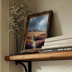May include: An ornate framed landscape painting depicts a path leading to a small house under a stormy sky. The painting is propped on a wooden shelf with books and a decorative floral arrangement.