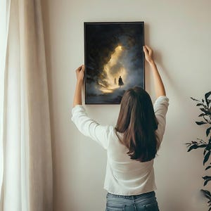 May include: A framed art print depicting a dark, atmospheric scene with figures in a luminous opening. The artwork, in a black frame, is being held up against a white wall. The image features a dramatic interplay of light and shadow.