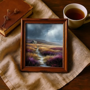 May include: A framed landscape painting depicts a path leading to a small house under a cloudy sky. The artwork is in a wooden frame, resting on a beige cloth. A cup of tea and a book are also present, creating a cozy, rustic scene.