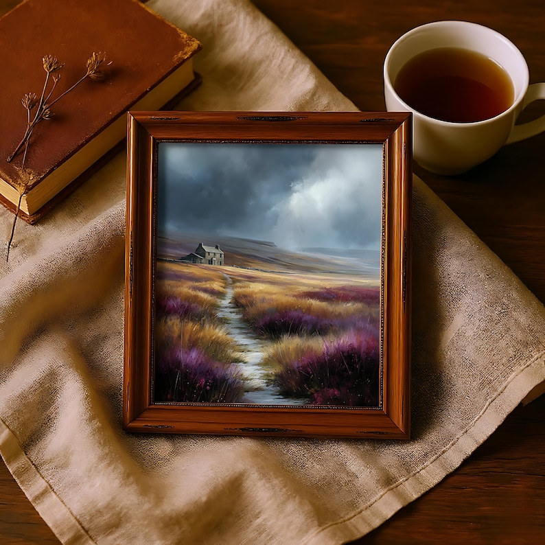 May include: A framed landscape painting depicts a path leading to a small house under a cloudy sky. The artwork is in a wooden frame, resting on a beige cloth. A cup of tea and a book are also present, creating a cozy, rustic scene.