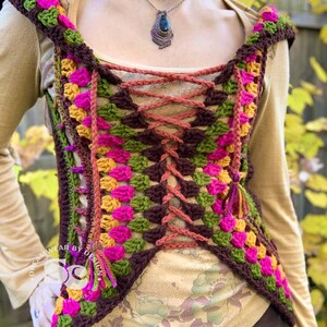 May include: A crocheted vest with a hooded neckline, featuring a colorful geometric pattern in shades of pink, green, brown, and yellow. The vest has a lace-up front with brown cords.