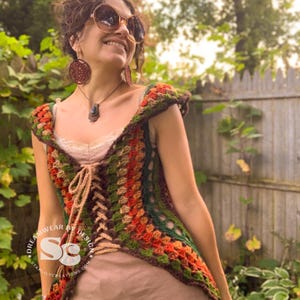 May include: A crocheted vest with a brown, green, orange, and beige color scheme. The vest has a lace-up front and a scalloped neckline. The vest is worn over a brown dress. The text "Dreamwear by Design" and "Starlilycreations.com" is visible on the vest.