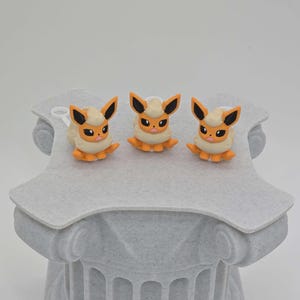 Puede incluir: Tres figuras de Eevee, cada una con una melena cremosa y esponjosa, cuerpo naranja y orejas con puntas negras. Las figuras se exhiben sobre un pedestal gris texturizado con un dise&ntilde;o de columna cl&aacute;sica. El fondo es blanco.