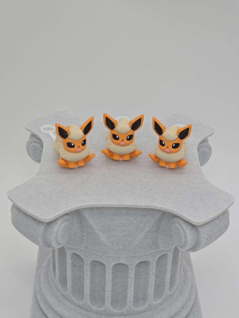 Puede incluir: Tres figuras de Eevee, cada una con una melena cremosa y esponjosa, cuerpo naranja y orejas con puntas negras. Las figuras se exhiben sobre un pedestal gris texturizado con un dise&ntilde;o de columna cl&aacute;sica. El fondo es blanco.