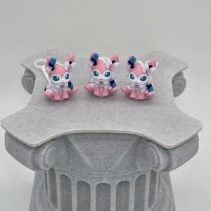 Puede incluir: Tres peque&ntilde;as y coloridas figuras de un personaje Sylveon se exhiben sobre un pedestal decorativo gris. Las figuras presentan detalles en rosa, blanco y azul. El pedestal tiene un dise&ntilde;o de columna cl&aacute;sica.