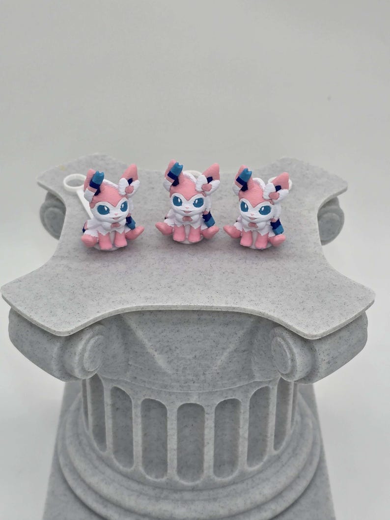 Puede incluir: Tres peque&ntilde;as y coloridas figuras de un personaje Sylveon se exhiben sobre un pedestal decorativo gris. Las figuras presentan detalles en rosa, blanco y azul. El pedestal tiene un dise&ntilde;o de columna cl&aacute;sica.