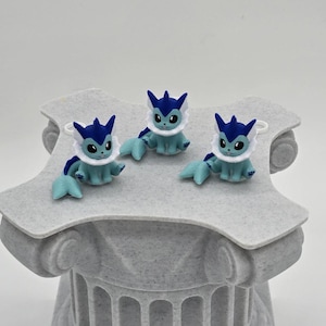 Puede incluir: Tres peque&ntilde;as figuras azules y blancas de una criatura de dibujos animados con ojos grandes, sentadas sobre una columna decorativa gris y texturizada. Las figuras tienen aletas y orejas azules, y detalles blancos similares a piel. La columna tiene un dise&ntilde;o cl&aacute;sico.