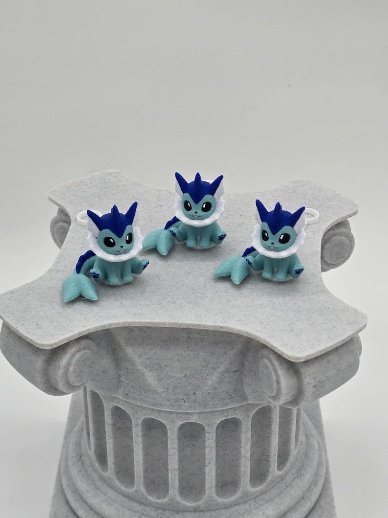 Puede incluir: Tres peque&ntilde;as figuras azules y blancas de una criatura de dibujos animados con ojos grandes, sentadas sobre una columna decorativa gris y texturizada. Las figuras tienen aletas y orejas azules, y detalles blancos similares a piel. La columna tiene un dise&ntilde;o cl&aacute;sico.