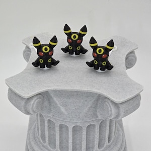 Puede incluir: Tres figuras negras de Umbreon con detalles amarillos y ojos rojos se exhiben sobre una columna decorativa gris texturizada. Las figuras est&aacute;n en posici&oacute;n sentada. La columna tiene un dise&ntilde;o cl&aacute;sico.