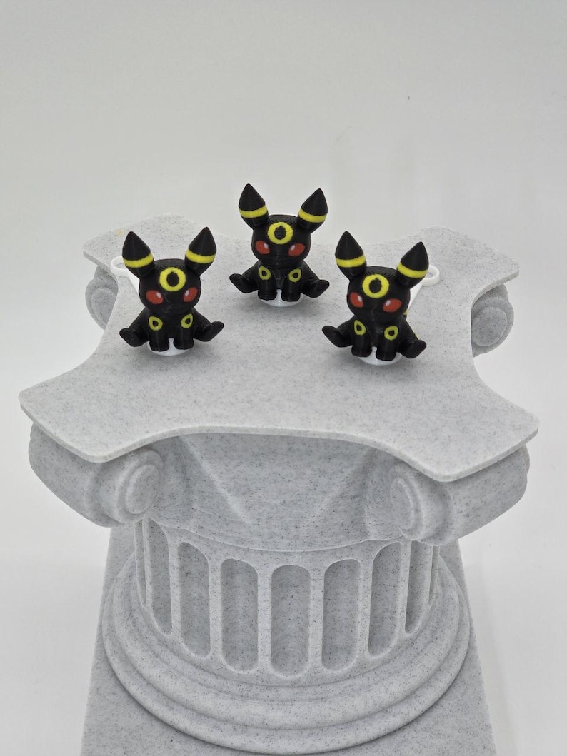 Puede incluir: Tres figuras negras de Umbreon con detalles amarillos y ojos rojos se exhiben sobre una columna decorativa gris texturizada. Las figuras est&aacute;n en posici&oacute;n sentada. La columna tiene un dise&ntilde;o cl&aacute;sico.