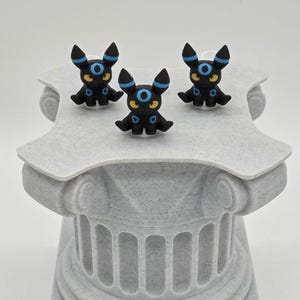 Puede incluir: Tres figuras negras de Umbreon con detalles azules y amarillos se exhiben sobre un pedestal gris texturizado. Las figuras tienen orejas grandes y ojos azules circulares. El pedestal tiene un dise&ntilde;o de columna cl&aacute;sica.