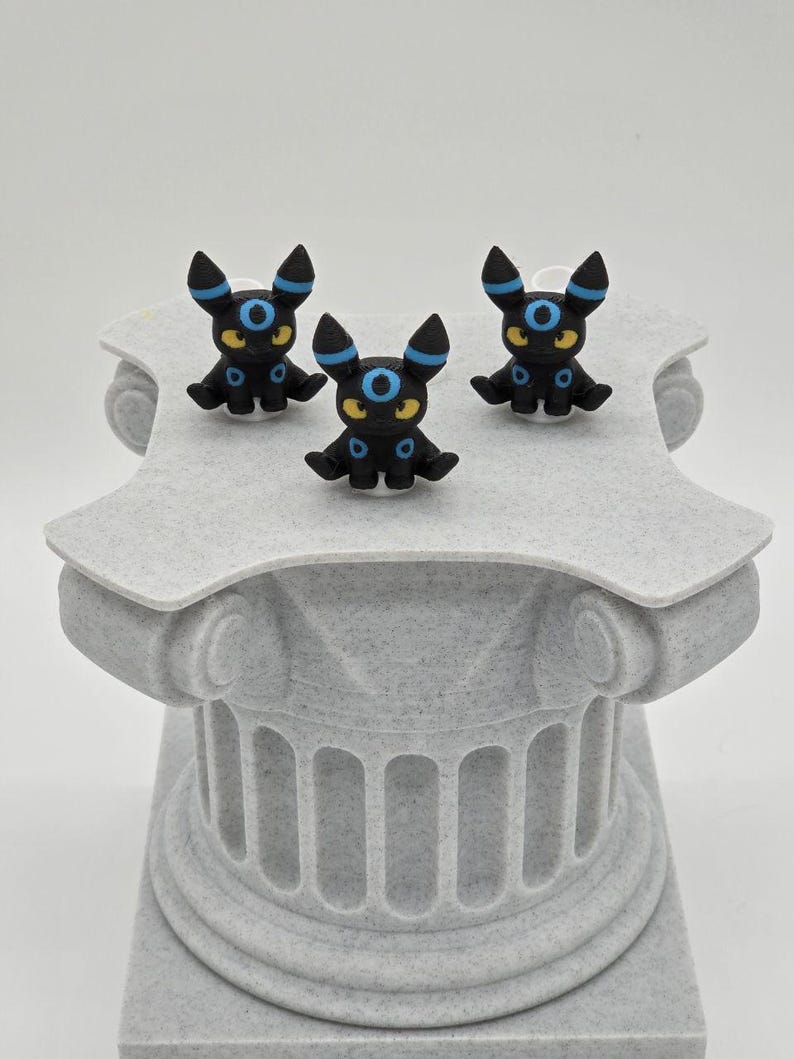 Puede incluir: Tres figuras negras de Umbreon con detalles azules y amarillos se exhiben sobre un pedestal gris texturizado. Las figuras tienen orejas grandes y ojos azules circulares. El pedestal tiene un dise&ntilde;o de columna cl&aacute;sica.