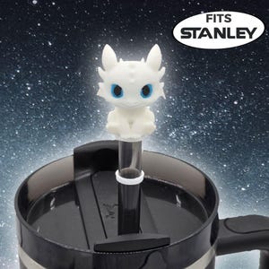 May include: A white dragon figure with blue eyes sits atop a clear straw, designed to fit a Stanley tumbler. The dragon has pointed ears and a textured surface. The tumbler is black with a black lid and handle.