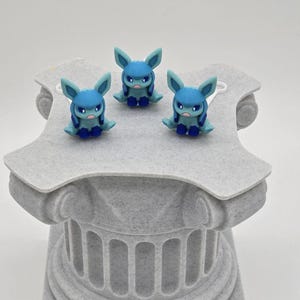 Puede incluir: Tres figuras azules con orejas grandes y expresiones severas se exhiben sobre un pedestal gris texturizado. Las figuras tienen cuerpos, orejas y acentos azules. El pedestal es de dise&ntilde;o de columna cl&aacute;sica.