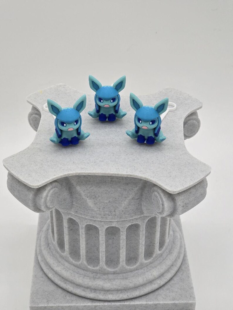 Puede incluir: Tres figuras azules con orejas grandes y expresiones severas se exhiben sobre un pedestal gris texturizado. Las figuras tienen cuerpos, orejas y acentos azules. El pedestal es de dise&ntilde;o de columna cl&aacute;sica.