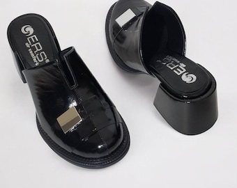 May include: Pair of black patent leather heeled mules with a silver buckle detail. The shoes have a low block heel and a closed toe.