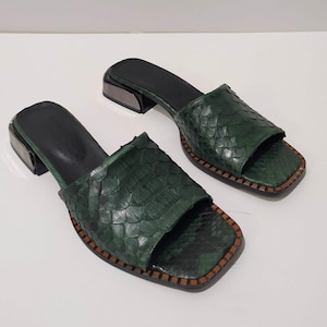 Könnte beinhalten: Ein Paar grüne Schlangenledersandalen mit silbernem Metall-Absatz. Die Sandalen haben eine quadratische Zehenpartie und einen niedrigen Absatz.