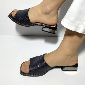 Könnte beinhalten: Ein Paar schwarze Sandalen mit Absatz und Schlangenhautmuster. Die Sandalen haben eine quadratische Zehenpartie und einen silbernen Absatz.