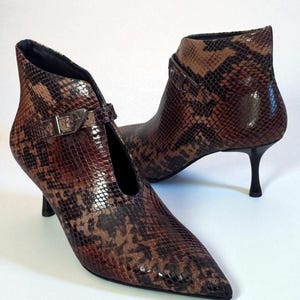 Könnte beinhalten: Ein Paar braun-beige Stiefeletten mit Schlangenmuster, spitzen Zehen und kleinen, schwarzen Stiletto-Absätzen. Jeder Stiefel hat eine silberne Schnalle an der Seite. Die Stiefel stehen vor einem weißen Hintergrund.