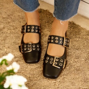 May include: Black leather shoes with gold buckles and eyelets. The shoes have two straps with buckles and are worn with denim jeans.