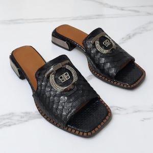 Pode incluir: Um par de sandálias de salto alto de couro preto com padrão de pele de cobra. As sandálias têm uma fivela grande de prata com strass na frente.