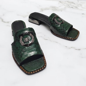 May include: A pair of green leather sandals with a snakeskin pattern and a silver buckle. The sandals have a low heel and an open toe.