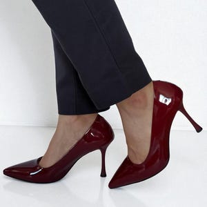 May include: Burgundy patent leather stiletto heels with pointed toes. The high heels have a slender, curved heel design. The shoes are paired with dark navy blue trousers. The shoes are a classic style.