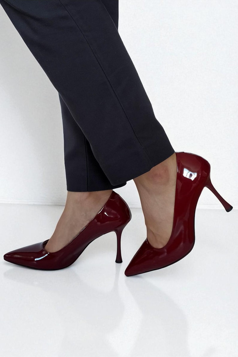 May include: Burgundy patent leather stiletto heels with pointed toes. The high heels have a slender, curved heel design. The shoes are paired with dark navy blue trousers. The shoes are a classic style.